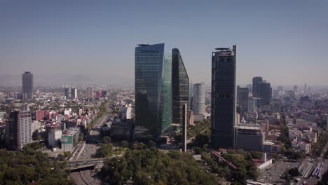Camera-shot-descending-from-skyscrapers-on-Paseo-de-la-Reforma-Avenue-in-Mexico-City-on-a-sunny-and-clear-day,-next-to-it-is-a-large-avenue-with-several-cars-circulating
