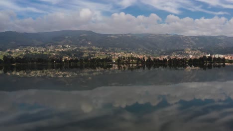 The-camera-flies-over-the-calm-reflective-waters-of-lake-Sochagota,-revealing-blue-skies-and-white-clouds-over-the-picturesque-town-of-Paipa,-in-Colombia,-South-America