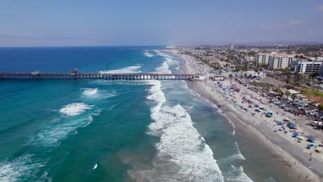 Drone-Flying-Over-Oceanside-Beach-Towards-Pier-and-Beachfront-Resorts