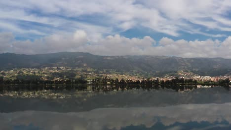 Fly-over-a-lake-that-reflects-the-blue-sky,-white-clouds,-and-picturesque-hills-of-Paipa,-Colombia,-like-a-natural-mirror