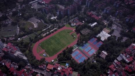 Drone-shot-of-sports-field-with-tennis-courts,-running-track,-soccer-fields,-south-of-Mexico-City,-the-complex-is-surrounded-by-residential-buildings-with-red-roofs