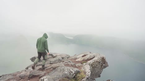 Man-Hiker-In-Hood-Walking-Towards-The-Cliff-Ledge-Looking-Down-The-Mountain-On-A-Foggy-Day-At-Segla,-Senja,-Norway