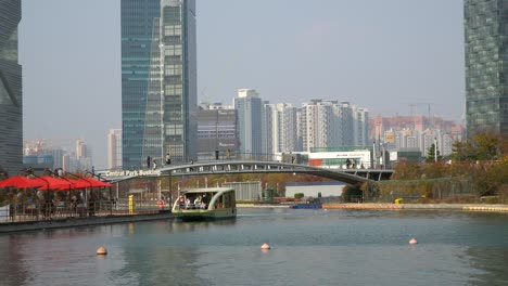 Songdo-Central-Park---People-Travel-by-Water-Taxi-Boat-on-a-Lake,-Travelers-Crossing-a-Footbridge-Enjoying-Futuristic-Moden-Urban-Skyline