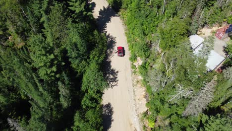 Rising-Aerial-Shot-of-Red-Vehicle-on-Dirt-Road-Surrounded-by-Green-Forest-on-Hot-Summer-Day