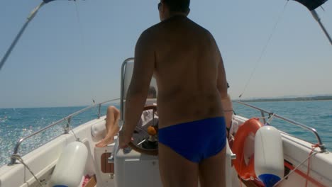Back-view-of-man-with-blue-swimwear-driving-fast-motorboat-steering-wheel-with-people-sunbathing-on-bow