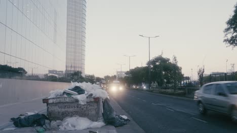 Stationary-shot,-huge-dumpster-in-front-of-Paseo-del-Bajo-complex-at-downtown-Buenos-Aires,-over-filled-with-rubbish-and-junk-due-to-municipal-mismanagement,-traffic-crossing-on-Eduardo-Madero-avenue