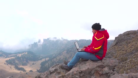 digital-nomad,-young-latin-man-working-on-his-laptop-sitting-on-a-rock-in-the-mountain,-backpacking