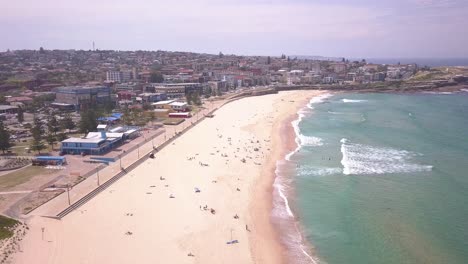 Reverse-aerial-flight-over-sandy-beach-at-the-eastern-suburb-Maroubra-town-center-and-blue-transparent-ocean-water