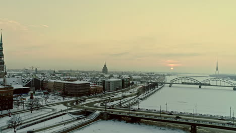 Aerial-drone-shot-of-busy-road-and-railway-bridge-over-the-frozen-river-and-city-of-Riga-by-the-banks-of-the-river-in-Latvia-2021-on-a-cold-wintry-sunset