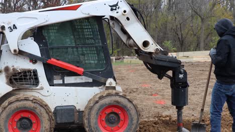 Construction-worker-uses-shovel-to-remove-loose-dirt-while-skid-steer-loader-with-hydraulic-auger-removes-auger-from-hole-and-drives-forward-to-remove-dirt-from-auger