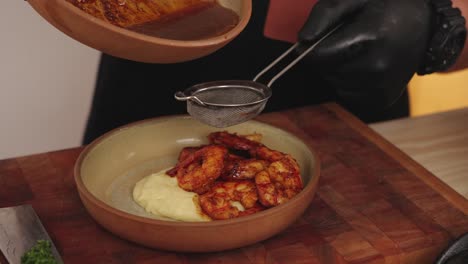 Faceless-chef-using-a-food-strainer-straining-the-sauce,-adding-the-liquid-into-a-plated-seafood-gourmet,-shrimps-with-mashed-potato-dish