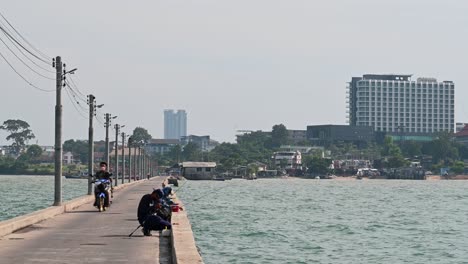 A-man-preparing-his-fishing-rod-while-a-motorcycle-passes-by-and-the-city-of-Pattaya-in-the-background-at-the-Pattaya-Fishing-Dock,-Chonburi,-Thailand