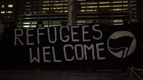 Protestors-hold-up-a-large-black-anti-fascist-banner-that-says-“Refugees-Welcome”-outside-the-UK-Home-Office-at-night-on-a-protest-for-refugee-rights