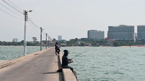 Un-Hombre-Pescando-Tambaleándose-Y-Levantando-Su-Caña-Con-Otros-En-El-Fondo-Y-La-Hermosa-Ciudad-De-Pattaya,-Muelle-De-Pesca-De-Pattaya,-Chonburi,-Tailandia