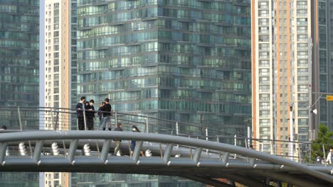 The-walking-or-pedestrian-bridge-to-Songdo-Central-Park-in-Incheon,-South-Korea,-people-wearing-face-masks-and-a-modern,-glass-skyscraper-in-the-background
