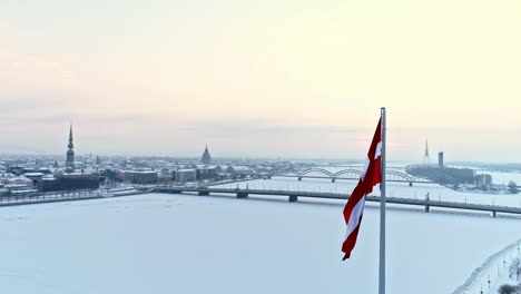 Latvian-flag-streams-in-breeze-next-to-frozen-Daugava-river,-Riga