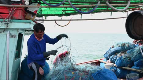 Mending-the-net-while-smoking-then-smiles-at-the-camera-as-the-boat-moves-with-the-waves,-Pattaya-Fishing-Dock,-Chonburi,-Thailand