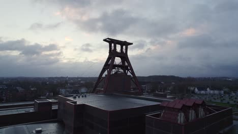 Zeche-Zollverein-shaft-12-winding-tower,-backlit-against-cloudy-dawn-sky,-drone-zoom-in