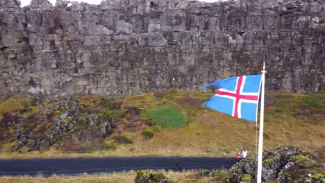 Icelandic-flag-on-a-windy-day-at-Thingvellir-National-Park,-Tourist-walking-around-Thingvellir-National-Park,-Iceland
