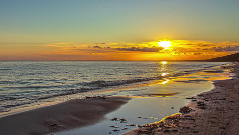 Time-lapse-of-vibrant-sunrise-along-coast,-small-waves-break-on-sandy-beach