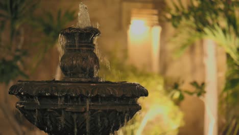 Water-Fountain-In-Courtyard-In-Evening-With-Bokeh-Wall-Lights-In-Background