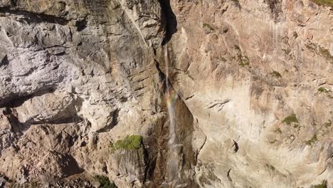 high-mountain-waterfall-from-the-rocky-wall-with-rainbow-colors,-aerial-pullback-shot