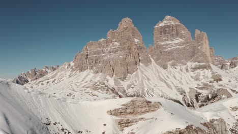 Hiker-walking-along-steep-snowy-ridge-line-with-Tre-Cime-dolomites-mountains-in-the-background