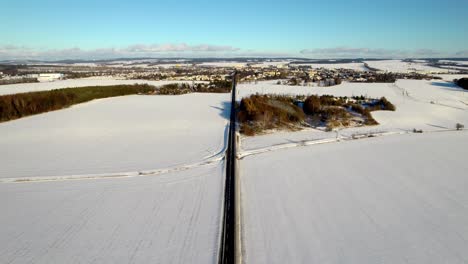 Aerial-view-of-a-straight-road-leading-to-the-city-as-cars-pass-through-the-snowy-rural-landscape