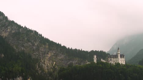 Aerial-dolly-in-of-dramatic-Neuschwanstein-Castle-on-a-hillside-surrounded-by-a-dense-green-pine-forest-on-a-foggy-overcast-day,-Bavaria,-Germany