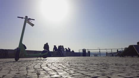 E-Scooter-Standing-On-Cobbled-Stones-With-Silhouette-Of-Tourists-Sitting-On-Wall-In-background-In-Lisbon