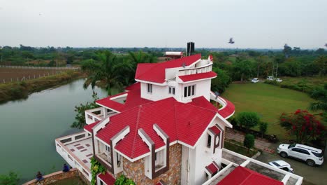 Black-birds-flying-in-a-flock,-in-rotation-to-a-luxurious-vacation-home,-with-an-aerial-drone-shot,-and-a-background-of-lake-and-palm-trees-with-a-feeling-of-relaxation-in-Vadodara,-India