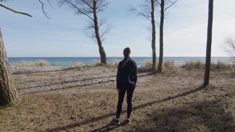 Young-healthy-fitness-woman-taking-exercise-break-looking-over-the-horizon-and-beach-from-a-glade-among-trees