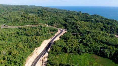 Blue-ocean-water-and-winding-road-under-construction-in-vibrant-forest-area-of-Indonesia,-aerial-view
