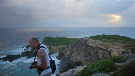 Middle-aged-male-tourist-with-a-camera-walks-into-a-viewpoint-to-take-a-photograph-of-waves-hitting-tall-rocky-cliffs-during-sunset