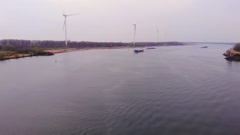 Aerial-View-Over-Oude-Maas-On-Overcast-Day-With-Still-Wind-Turbines-On-Rivers-Edge-And-Cargo-Ships-Approaching-In-Distance