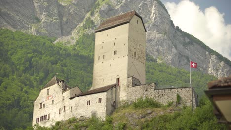 Low-angle-of-historical-Sargans-Castle-with-Swiss-flag-waving-on-a-hill-near-Mount-Gonzen-surrounded-by-dense-green-pine-forest,-Switzerland-Alps