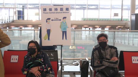 Passagiere-Sitzen-Neben-Einem-Schild,-Das-Die-Menschen-Daran-Erinnert,-Am-Internationalen-Flughafen-Chek-Lap-Kok-In-Hongkong,-China,-Soziale-Distanz-Einzuhalten