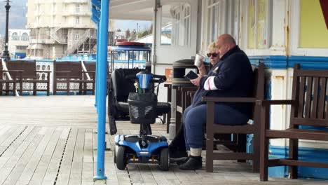 Elderly-retired-couple-with-mobility-scooter-relaxing-on-Welsh-seaside-pier-promenade-staycation