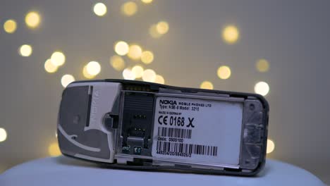 Graues-Nokia-Modell-3210-Handy-Ohne-Batterie-Dreht-Sich-Auf-Drehscheibe-Mit-Lichtern-Im-Hintergrund