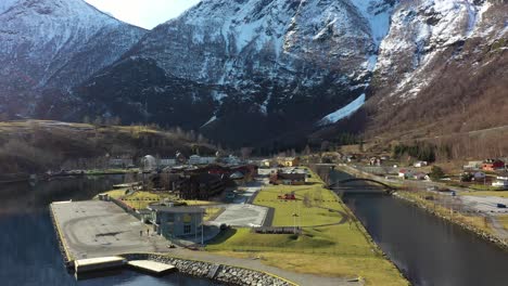 Beautiful-Flam-Norway---Backward-moving-aerial-over-clear-fjord-with-sun-and-mountain-reflections---From-closeup-of-town-and-cruise-quay-to-panoramic-overview-with-mountain-background