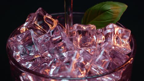 Static-shot-of-artistic-cocktail-with-light-red-or-pink-light-and-coloring-being-poured-over-ice-with-a-black-background-and-a-green-leaf-garnish