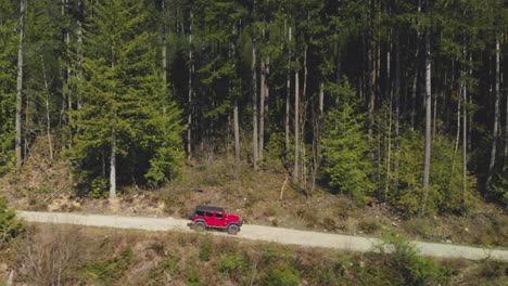 Red-jeep-SUV-off-road-vehicle-driving-on-dirt-logging-road-approaching-and-passing-dense-green-forest-view-in-background-Aerial-beside-trucking-tracking-as-truck-moves-to-right-accelerating-passed