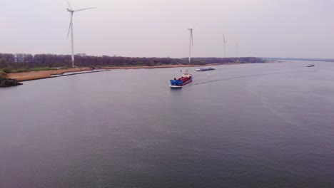 Aerial-View-Over-Oude-Maas-On-Overcast-Day-With-Still-Wind-Turbines-On-Rivers-Edge-And-General-Cargo-Ships-Approaching-In-Distance