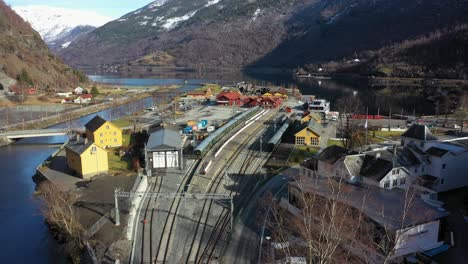 Flamsbanen-railway-station-in-Norway---Sunny-day-aerial-view-of-historic-museum-train-waiting-on-station-surrounded-by-river-and-tall-mountains---Slowly-moving-forward