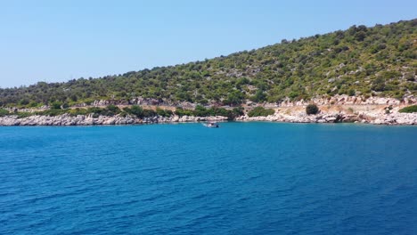 Wide-aerial-view-of-a-green-hill-next-to-the-tropical-blue-Mediterranean-Sea-in-Finike-Turkey-with-a-sailboat-anchored-in-the-water