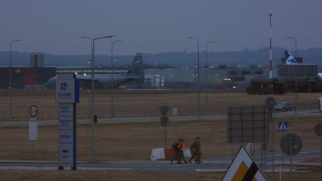A-military-cargo-plane-awaits-takeoff-in-the-distance,-four-airport-security-personnel-cross-a-road