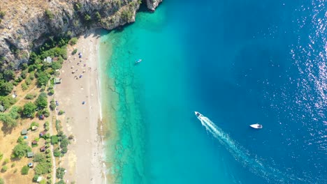 aerial-top-down-view-of-the-turquoise-blue-water-at-Butterfly-Valley-as-a-boat-approaches-a-white-sand-beach-in-Fethiye-Turkey-on-a-sunny-summer-day-surrounded-by-mountains
