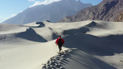 Drohne-Verfolgt-Einen-Jungen-Glücklichen-Kaukasischen-Männlichen-Touristen,-Der-In-Einer-Roten-Jacke-Unterwegs-Ist-Und-Tagsüber-Die-Spitze-Von-Sanddünen-In-Der-Kalten-Wüste-Von-Skardu-Pakistan-Mit-Einer-Bergkette-Im-Hintergrund-Läuft