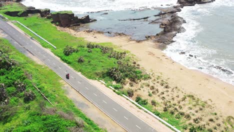 aerial-drone-view-of-two-people-on-a-two-wheeler-riding-on-a-well-paved-empty-road-with-a-sandy-beach-to-the-right,-white-waves-gently-crashing-on-the-shore-with-rocky-formations-and-greenery