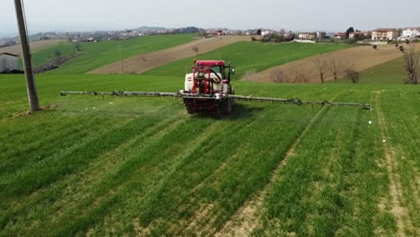 Aerial-view-of-red-farming-tractor-spraying-on-field-with-sprayer,-herbicides-and-pesticides-insecticide-to-the-green-field-plowed-land,-medium-shot